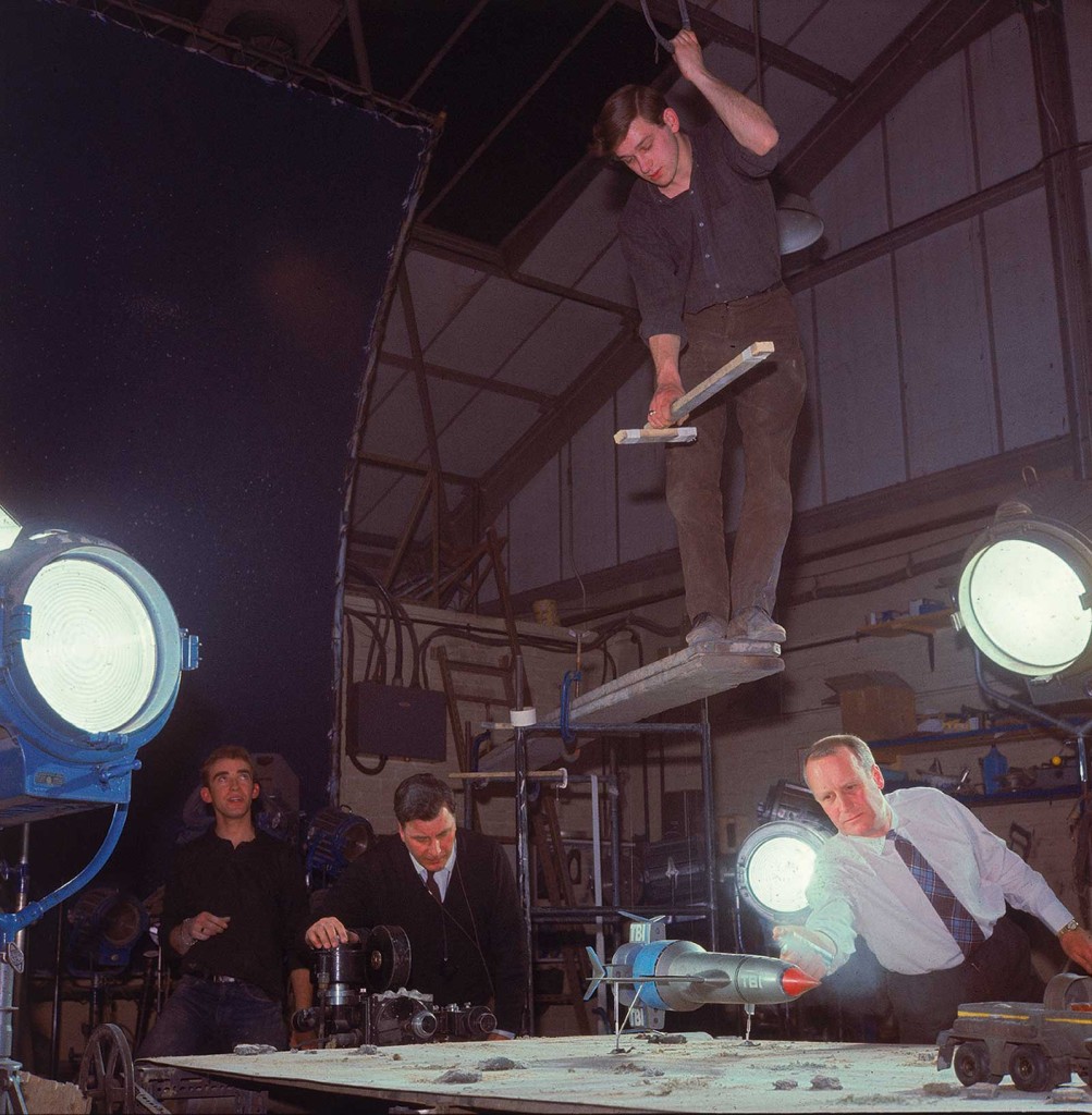 Special effects floor technician (far left) and special effects second unit lighting cameraman Harry Oakes (centre) preppare a scene for the episode Martian Invasion. Floor technician Peter Wragg balances on planks overhanging the set, ready to fly Thunderbird 1.