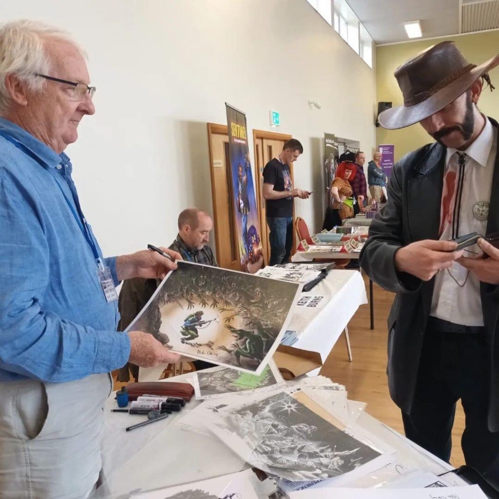 Mike Dorey with some of his art at Enniskillen Comic Art Festival, with Keith Burns drawing away in the background