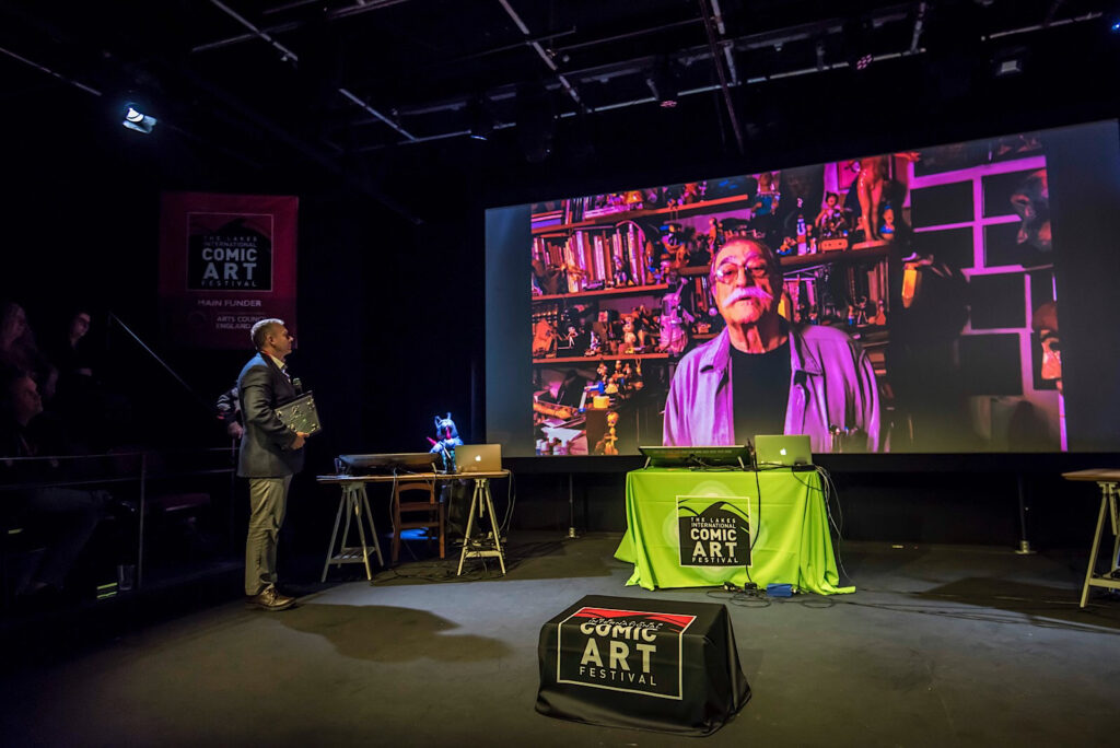 Tom Richmond announcing Posy Simmonds as the winner of the 2022 Sergio Aragonés Award at the Lakes International Comic Art Festival, Sergio on screen delivering a video message