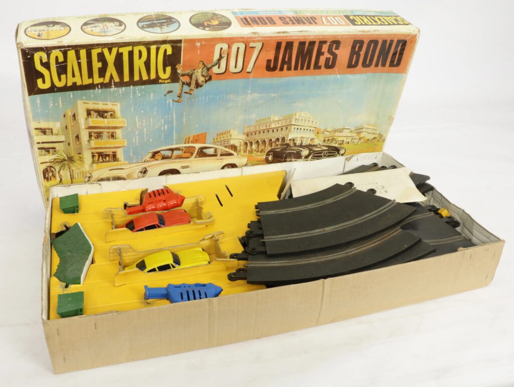Scalextric vintage 007 James Bond slot car racing set including curved track sections, red and blue hand controllers, incorrect slot cars in red and yellow, scenery components, and instructions