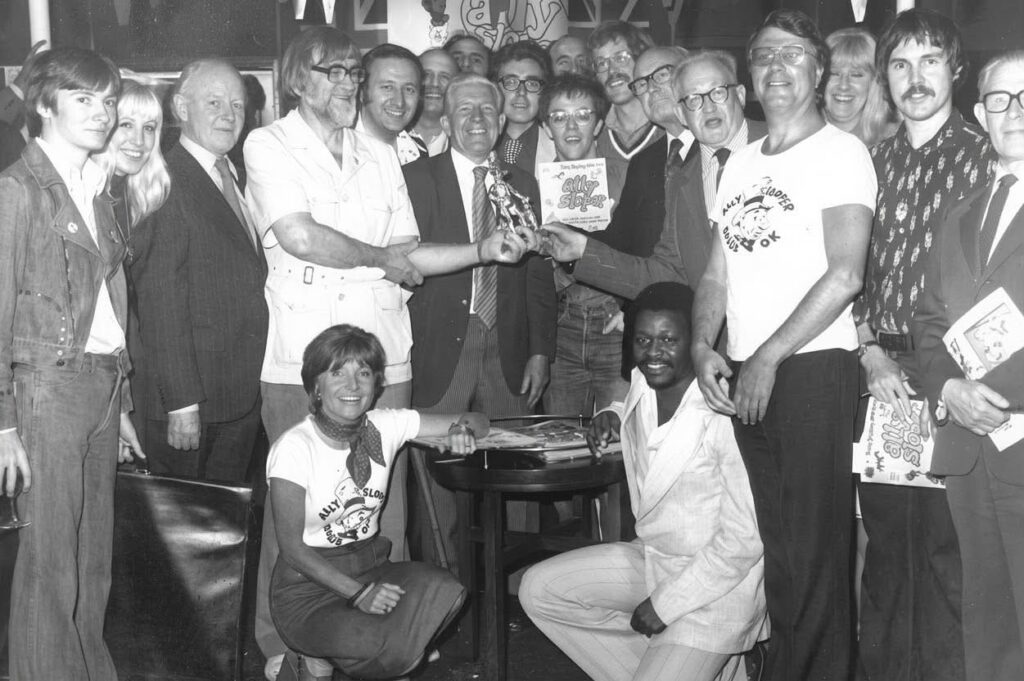 Britain's top cartoonists line up for a laugh at the Ally Sloper Launch Party on the  Tattersall Castle. Flanking Ted Ray, the only top wireless comedian never to appear in the weekly comic, Radio Fun are (strip credits listed here contemporary for 1976) Kevin O'Neill (then  late of "Legend Horror Classics"), Roland ("Come On Steve" Davies, Frank ("Dan Dare") Hampson, Alan ("Larri Read") Vince, Keith ("Hunter") Page, who came across this copy of the photograph in an old book, Hunt ("Dogman") Emerson, Tony Schofield, Walter ("Ally Sloper") Bell, Terry ("Morecambe and Wise") Wakefield, Denis ("Steadfast McStaunch") Gifford, Keith ("Jarl the Northman") Luck, and Fred ("Gremlins") Robinson. Almost in view are Tony ("Old Pals") Glynn, Eddie ("Kaptain Kween") McHenry, plus authors W.O.G ("man Behind Boys Fiction"), Lofts and Mary ("You're a Brick, Angela") Cadogan. Kneeling: Ann (Ms's Muse") Spano, and Ewan ("Miz Liz") Masters.