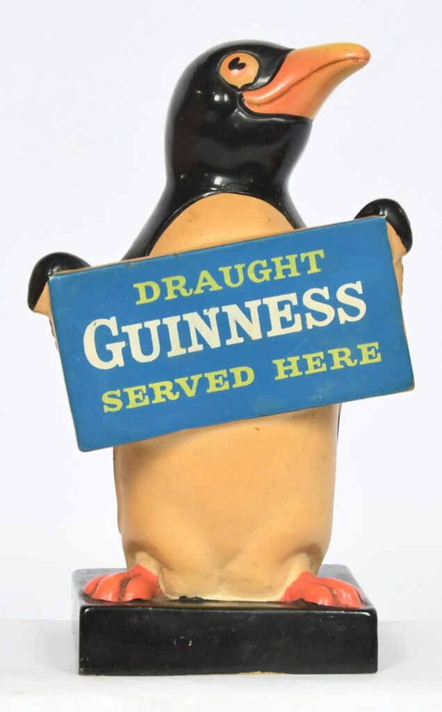 Charles E Tresise Ltd Guinness Penguin – a 1960s Original Rubberoid advertising bar figure, modelled standing on a black rectangular base and holding a blue sign reading 'DRAUGHT GUINNESS SERVED HERE'