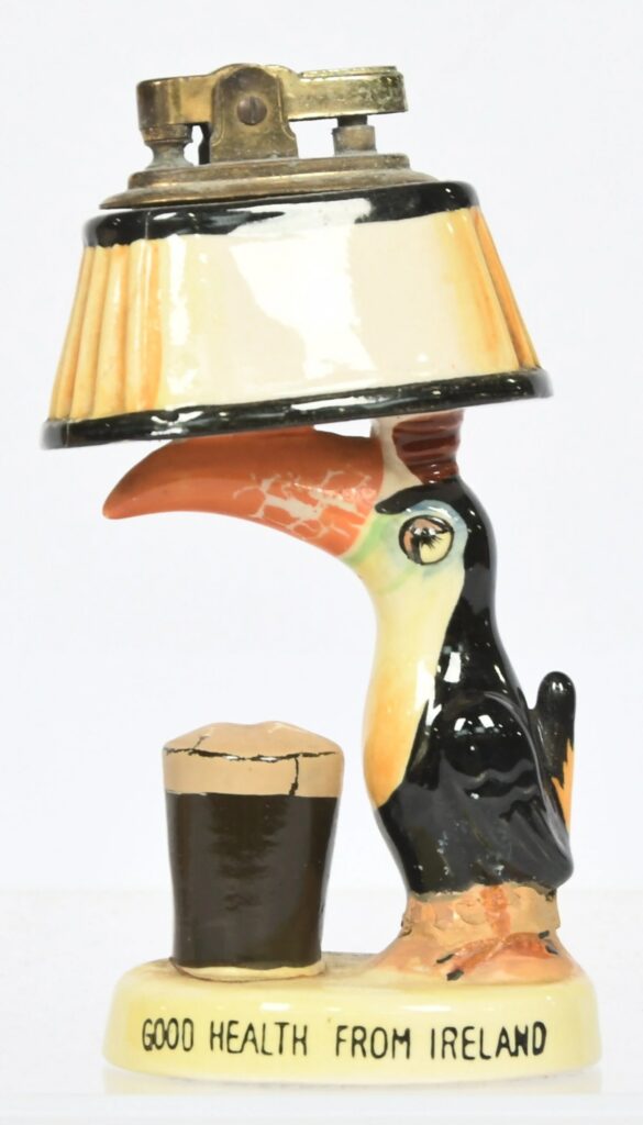 Guinness vintage ceramic novelty lighter, a figural table lighter modelled as a toucan with a lampshade on its beak standing beside a pint of stout on an oval plinth base, decorated to the front with the slogan "Good Health From Ireland". Complete with a brass-finish lighter mechanism inserted into the top